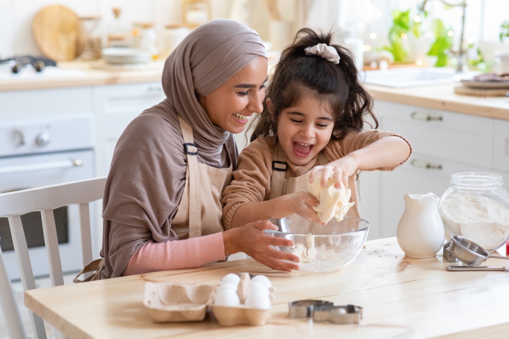 Une maman musulmane souriante, portant le hijab, et sa petite fille s'amusent à la maison, préparant des pâtisseries ensemble. Elles pétrissent la pâte à biscuits et prennent plaisir à cuisiner des plats faits maison.